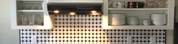 An image of a kitchen featuring dark work surfaces with cream kithcen cabinets and a monochrome tiled wall.