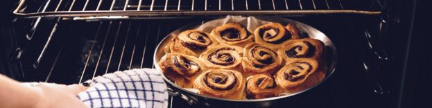 An image of a person using a tea towel to remove cinnamon buns from an electric oven